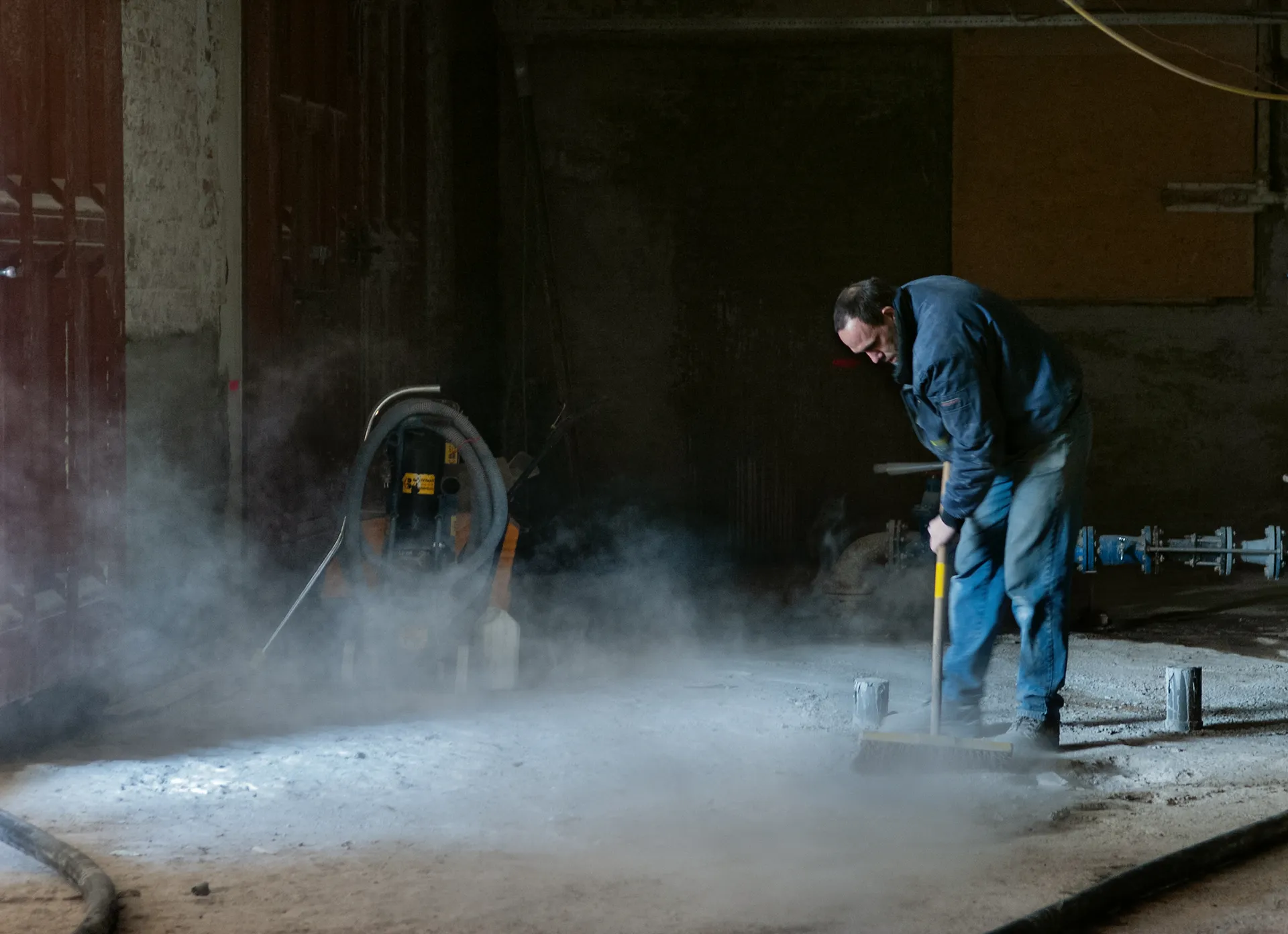 Een man in een blauwe overall gebruikt een schep om een stoffige vloer in een industriële omgeving schoon te maken.