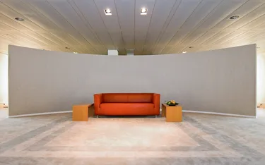 Een moderne wachtruimte in een kantoor met een comfortabele oranje bank, een houten tafeltje en een grote witte muur, voor een professionele uitstraling.