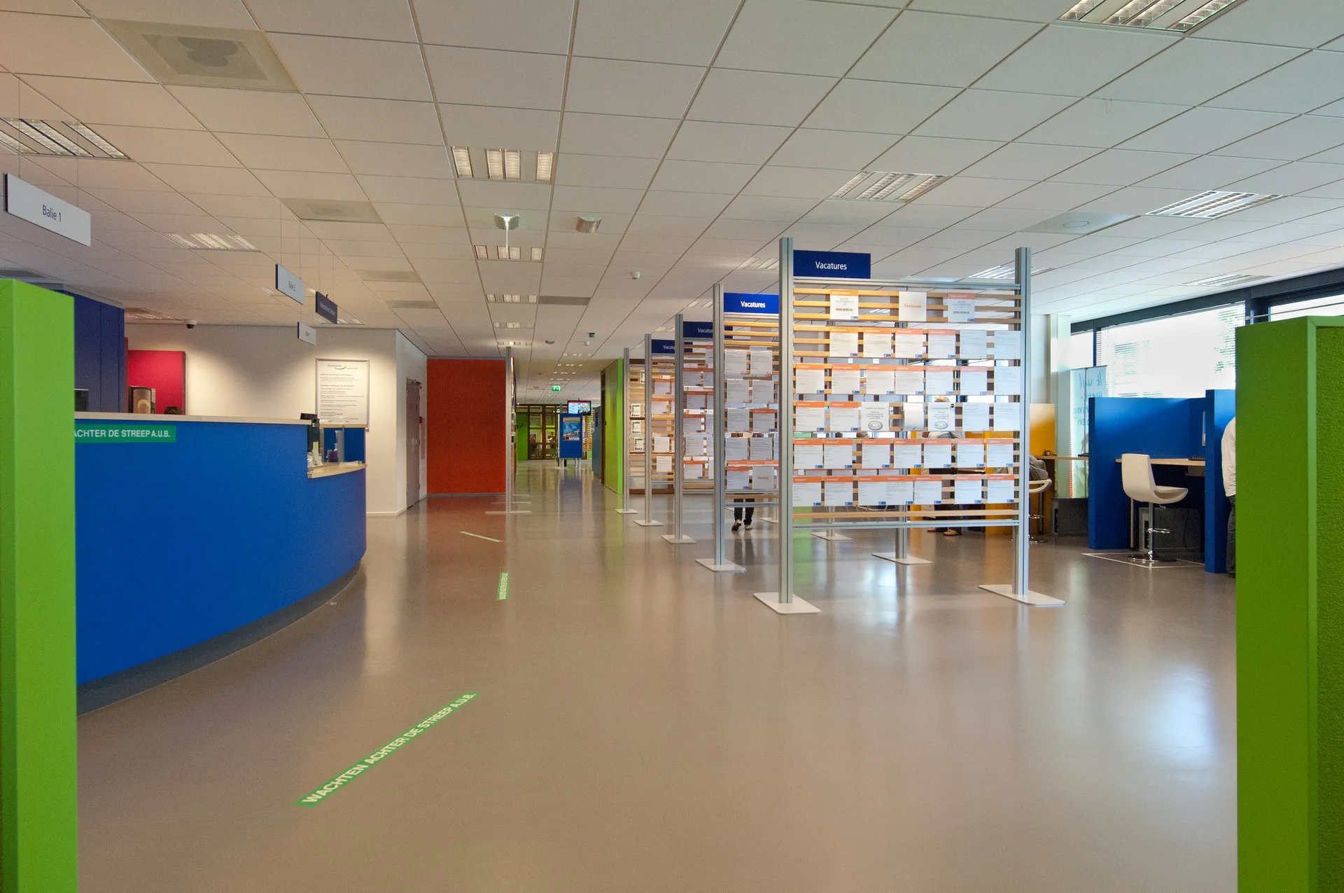 Een langgerekte, open kantoorruimte met een blauwe receptiebalie, groene en blauwe werkplekken, en een witte stoel op de achtergrond.