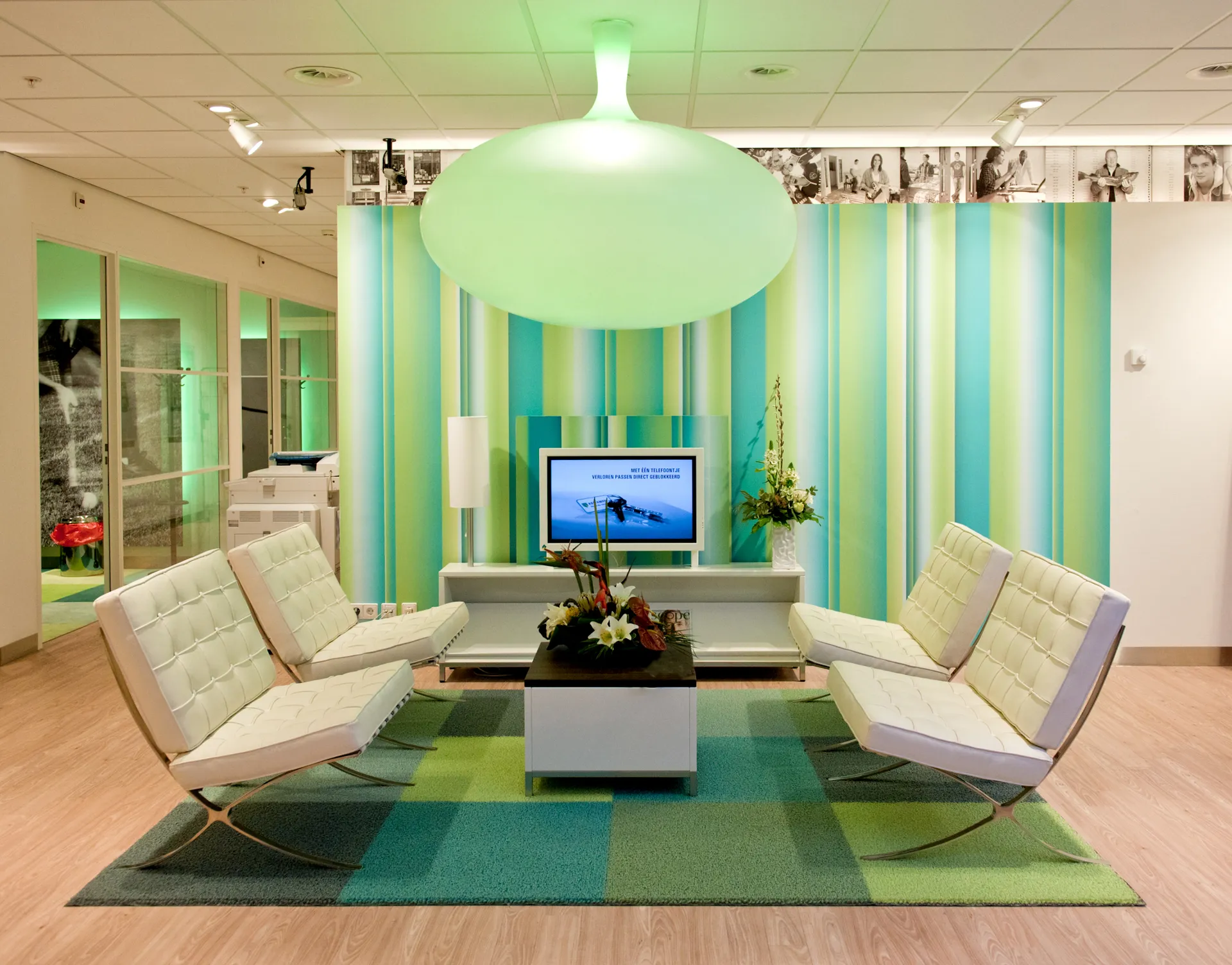 Een moderne wachtruimte met witte stoelen, een salontafel en een tv aan een muur met een groenblauw streeppatroon.