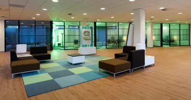 Een ruime, moderne wachtruimte met een geruit vloerkleed, comfortabele stoelen en een glazen wand die de ruimte scheidt van de rest van het kantoor.