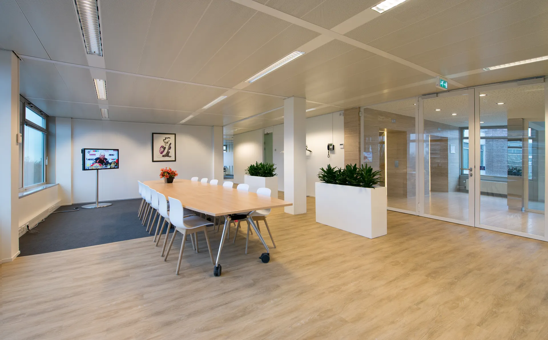 Een goed verlichte, moderne vergaderruimte in een kantoor met een grote tafel, witte stoelen en een tv-scherm aan de muur.