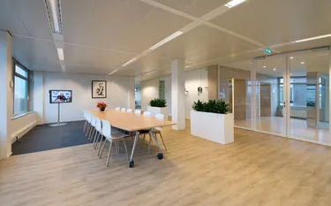 Een goed verlichte, moderne vergaderruimte in een kantoor met een grote tafel, witte stoelen en een tv-scherm aan de muur.