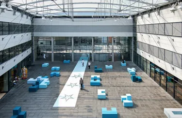 Een grote, open kantoorruimte met een hoog plafond en een centraal atrium, met een lange witte loopbrug met sterpatronen en blauwe zitelementen.