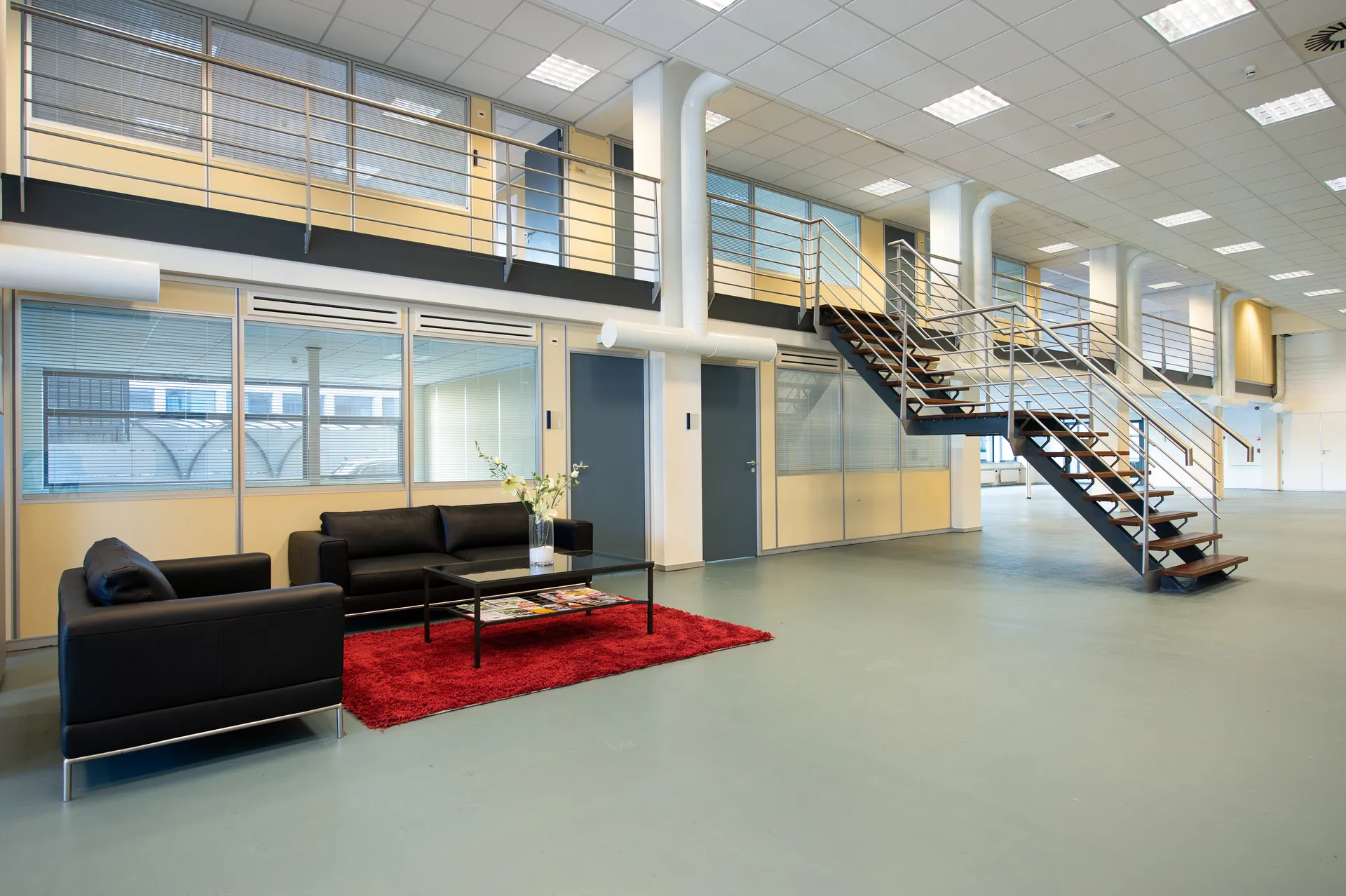 Een moderne kantoorruimte met een zwarte bank, rood vloerkleed en glazen salontafel, omgeven door grote ramen en een trap.