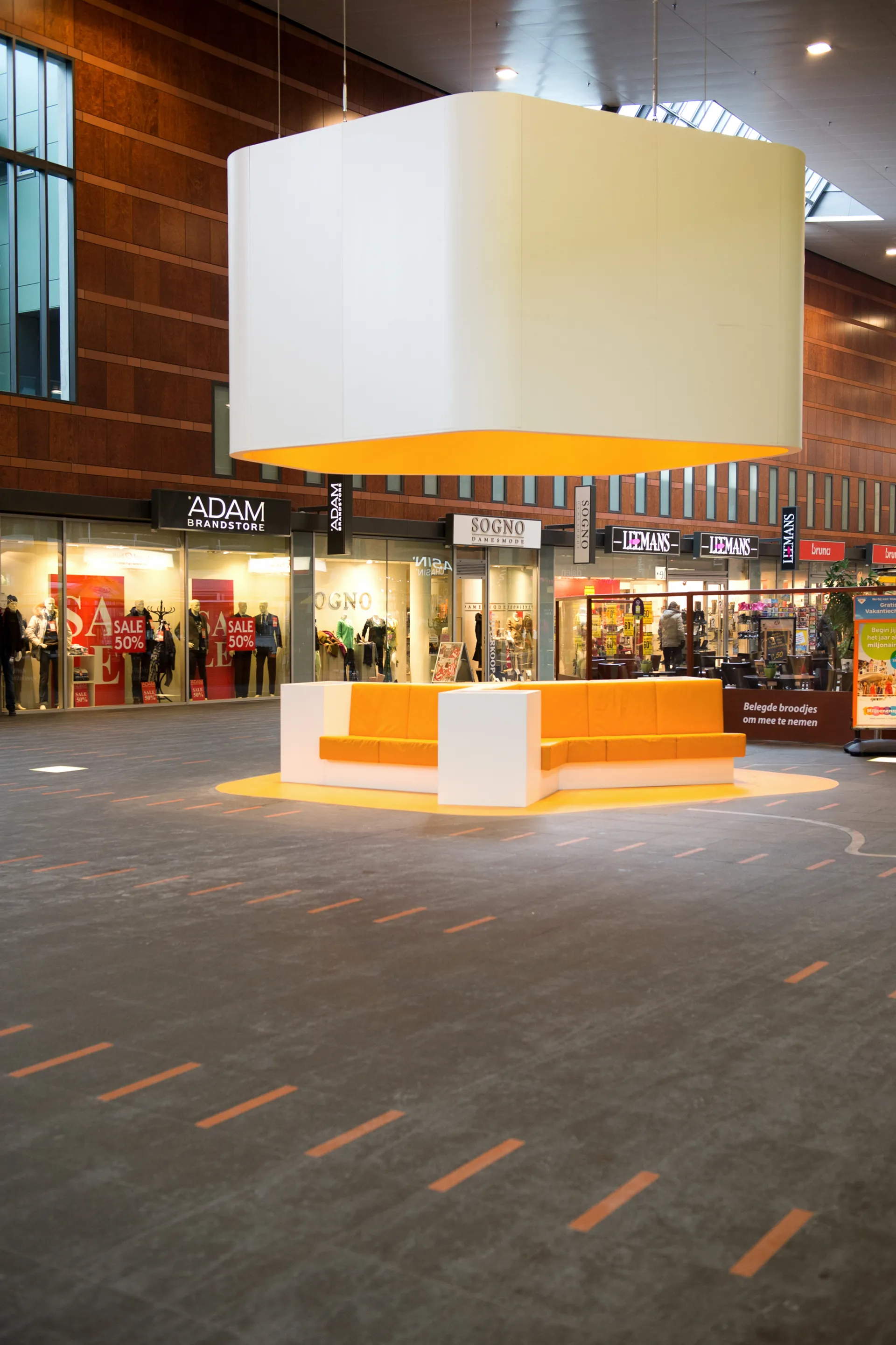 Een grote, open ruimte in een winkelcentrum met een modern jasje, met een groot wit-geel lichtornament en bankjes.