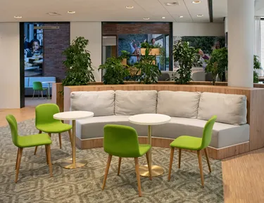 Een moderne pauzeruimte in een kantoor met een witte L-vormige bank, groene stoelen en kleine witte tafeltjes op een tapijt met patroon.