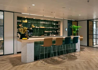 Een moderne bar binnen een kantoorruimte met een aanrecht, krukken en kasten, perfect voor informele overleggen.