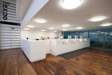 Een moderne receptieruimte in een kantoor met een witte balie, een strak design, glaswanden en een lichte houtkleurige vloer.