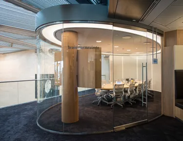 Een ruime overlegkamer met een grote glazen wand en een grote brede ronde vergadertafel vol stoelen, geschikt voor teamgesprekken.